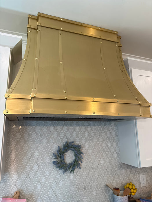 Custom brass range hood modern curved design with straps and rivets at RangeHoodMaster